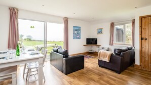 A spacious living area with a dining table, two sofas, a television, and large windows offering a view of the outdoors. - Puffin Lodge (Port Isaac)