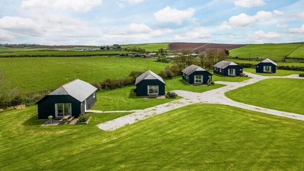 A rural landscape with several small, dark-colored cottages with pitched roofs, arranged in a cluster with a gravel path connecting them. - Puffin Lodge (Port Isaac)