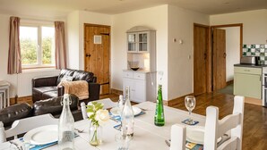 A dining area with a table set for two, a sofa, a cabinet, and a kitchen area with a toaster. - Puffin Lodge (Port Isaac)