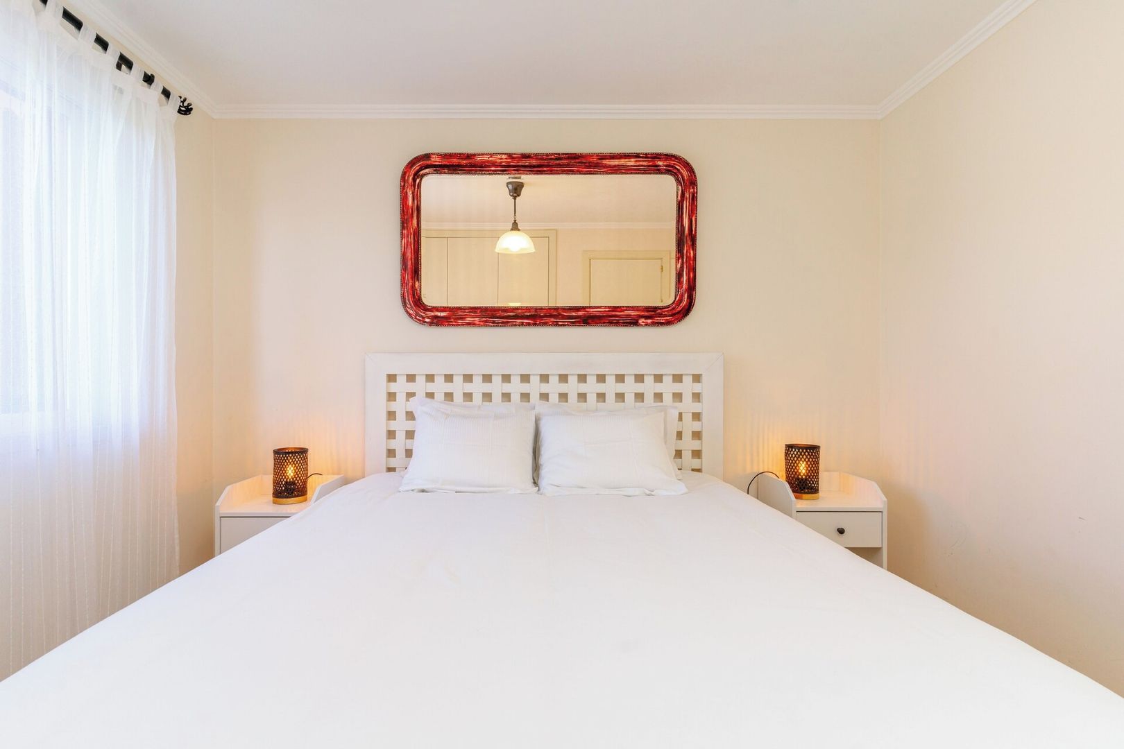 Een hotelkamer met een groot bed, twee nachtkastjes, een spiegel aan de muur en een raam met dunne gordijnen.