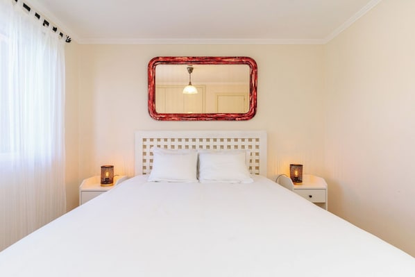 A hotel room with a large bed, two bedside tables, a wall-mounted mirror, and a window with sheer curtains.