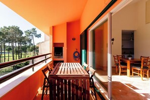 A balcony with a wooden table and chairs, a barbecue grill, and a view of trees.