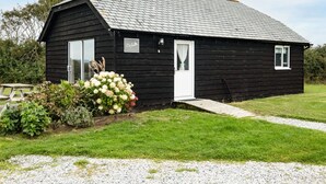 Una cabaña de madera negra con puerta y ventanas blancas, rodeada por un césped y un camino de grava.