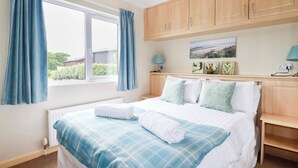 A bedroom with a bed, curtains, a window, a radiator, and a wall-mounted lamp. - Chi Prenn Byhan (Tintagel)