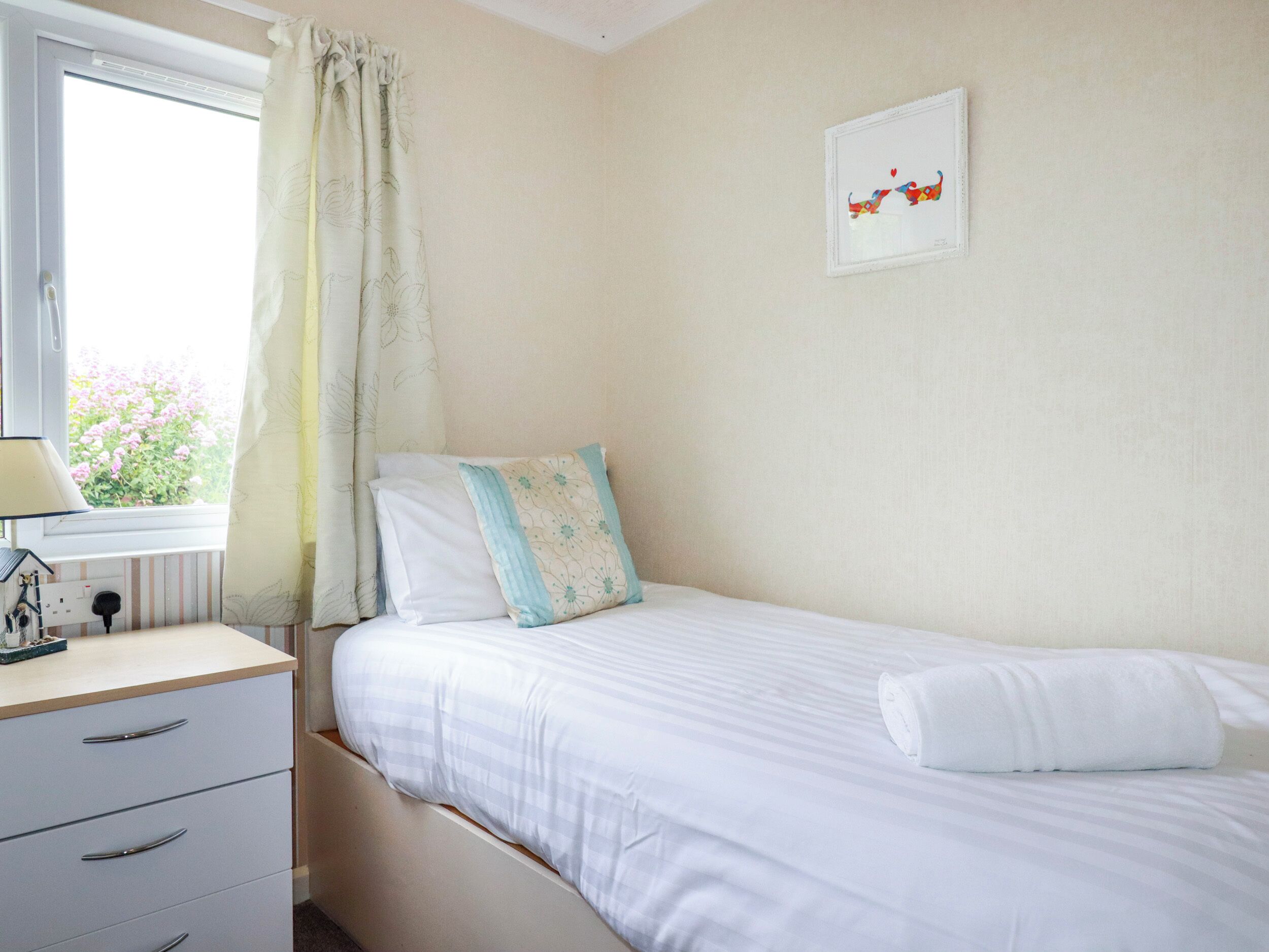 Quarto com uma cama de solteiro, mesa de cabeceira com abajur, janela com cortinas e um quadro na parede.