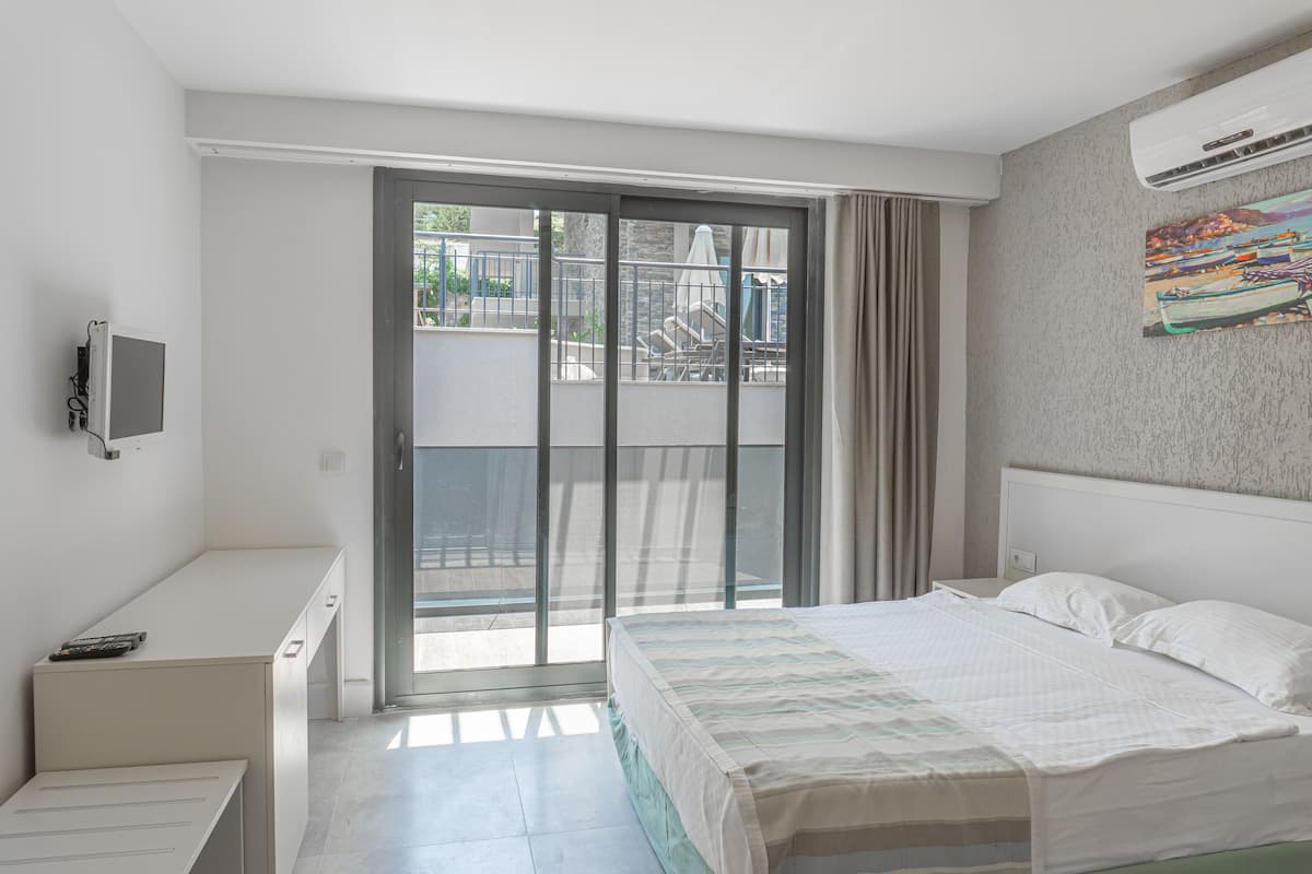 Un dormitorio con cama, escritorio, televisor y balcón con vistas al exterior.