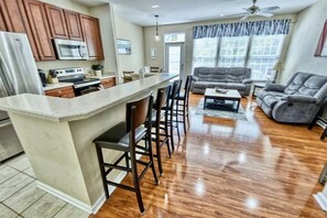 A modern kitchen with a central island, bar stools, a dining area with a sofa, and a large window with curtains.