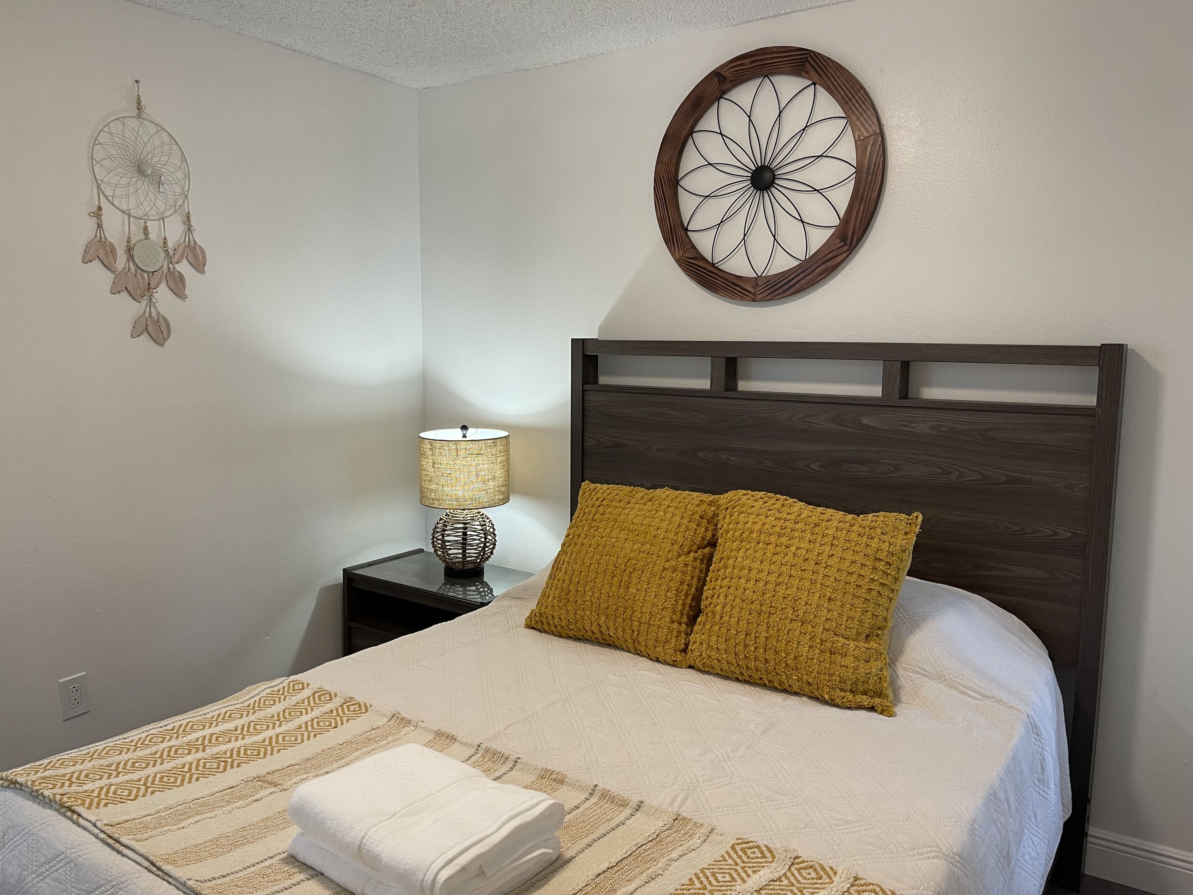 A neatly made bed with a patterned bedspread, two mustard-colored pillows, and a decorative dreamcatcher on the wall.