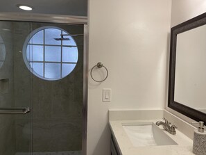 Ein Badezimmer mit Duschabtür aus Glas, einem runden Fenster, einem Handtuchhalter, einem Waschbecken und einem Spiegel.