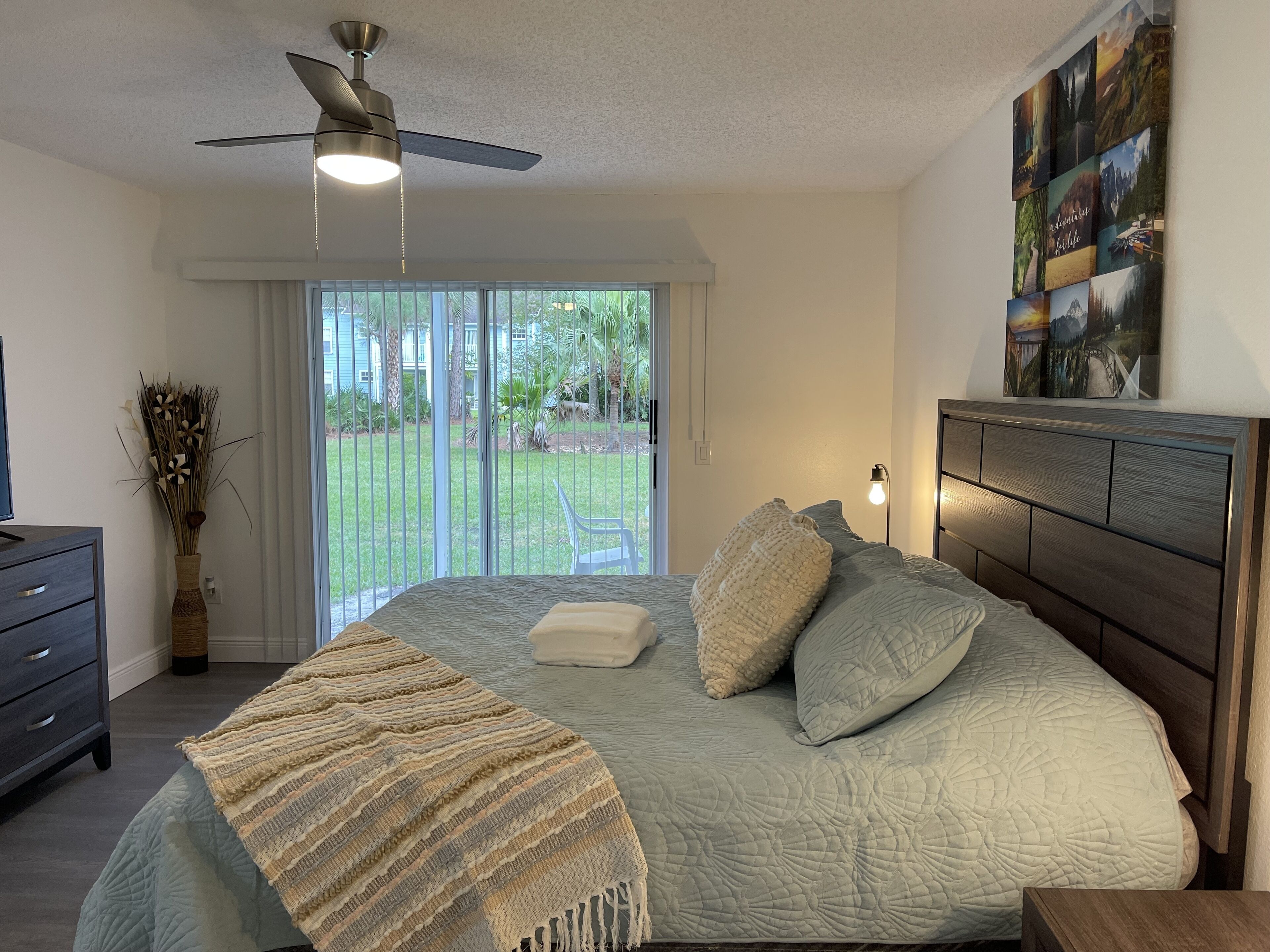 Quarto com cama, ventoinha no teto, mesa de cabeceira, cômoda e vista para o exterior através de uma porta de correr.