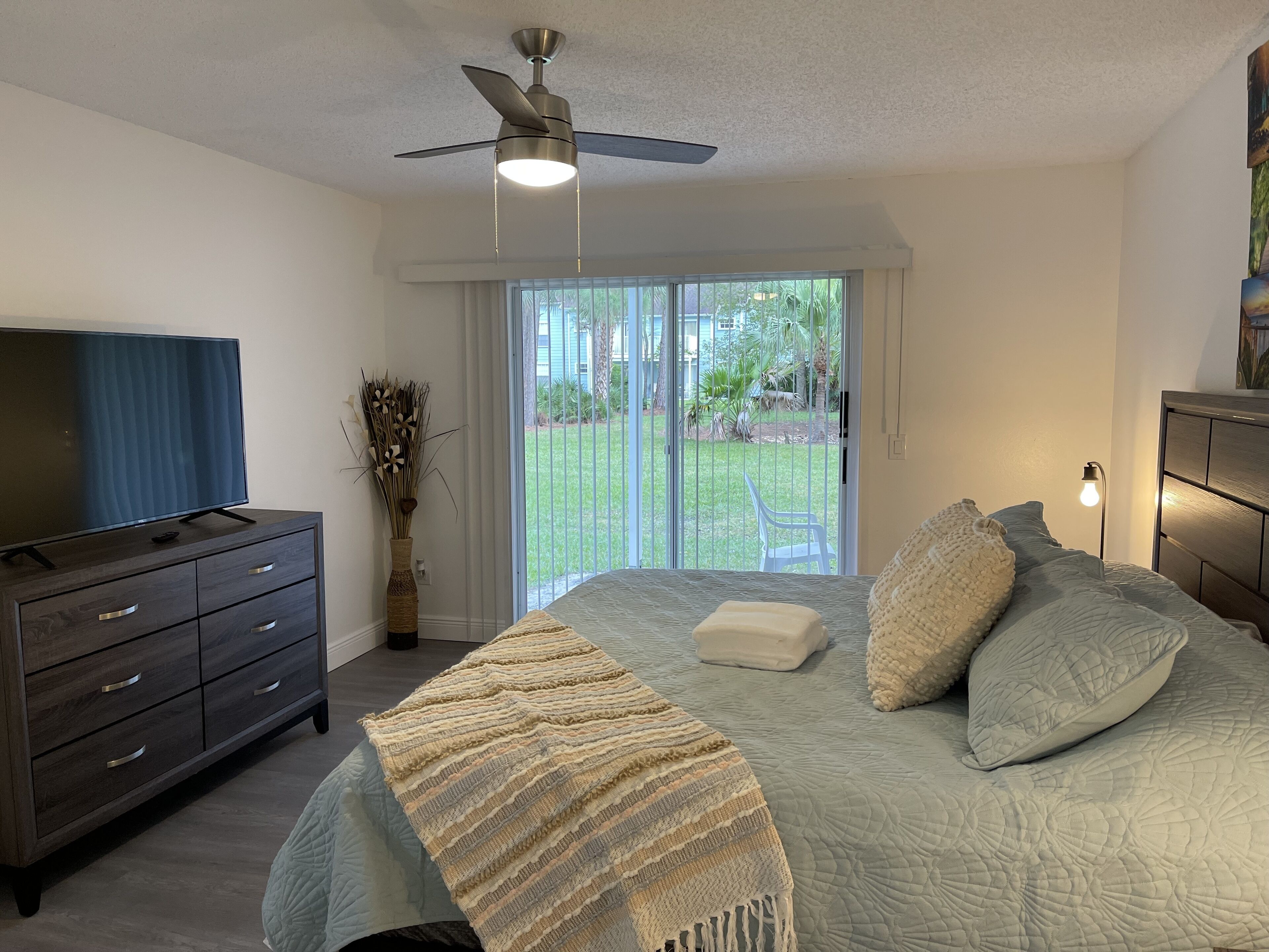 A bedroom with a bed, a dresser, a television, a ceiling fan, and a view of the outdoors through a sliding glass door.