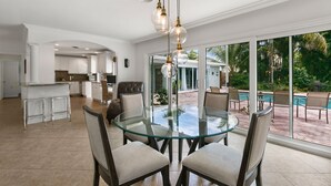 Un comedor amplio con una mesa y sillas de vidrio, una cocina al fondo y vistas a una piscina y follaje.
