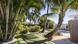 Un patio trasero con un campo de golfito, palmeras, una piscina y una cerca de madera.