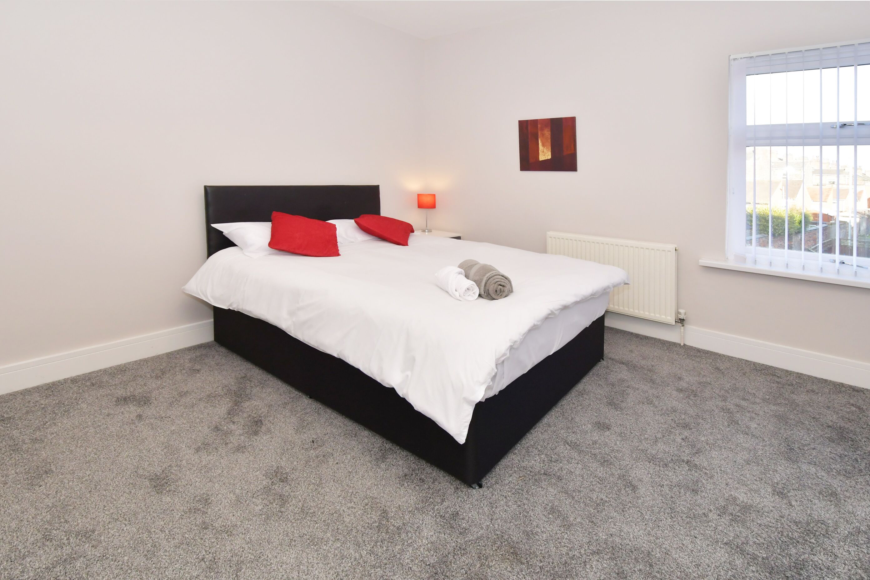 A neatly made bed with white and red pillows, a rolled towel, and a black headboard. There is a white radiator on the wall and a window with blinds.