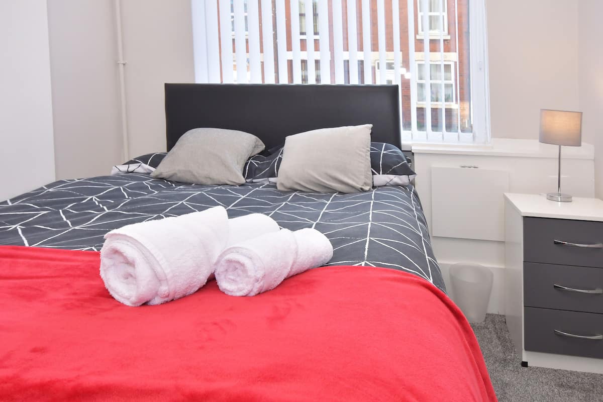 Una cama bien hecha con una colcha geométrica blanco y negro, dos almohadas y toallas blancas enrolladas sobre una manta roja.
