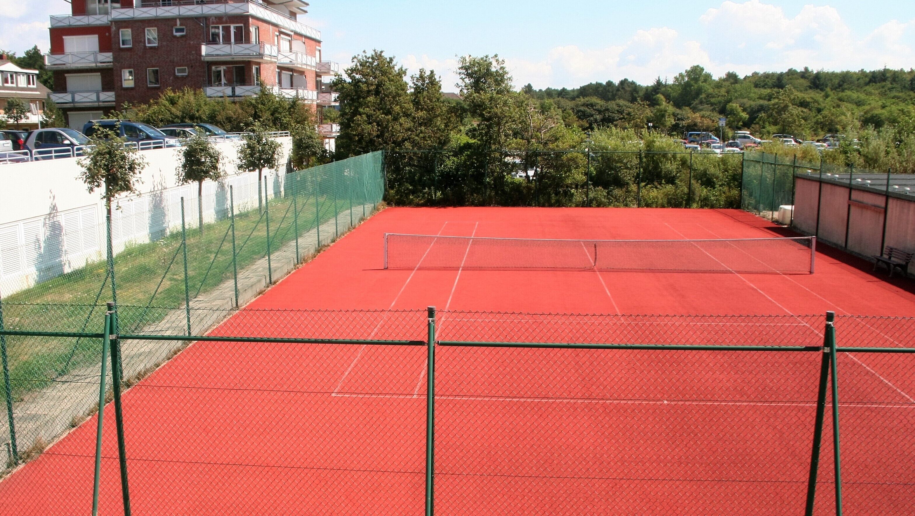 An outdoor tennis court with a red surface, surrounded by a green fence, and adjacent to a parking area with cars.