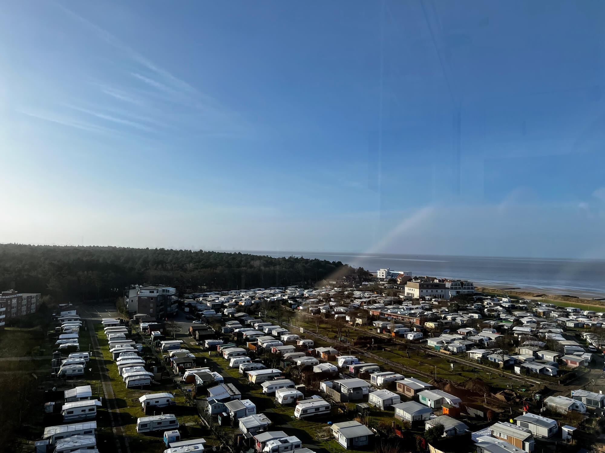 A large RV park with rows of RVs and trailers, a road, and a forested area in the background.