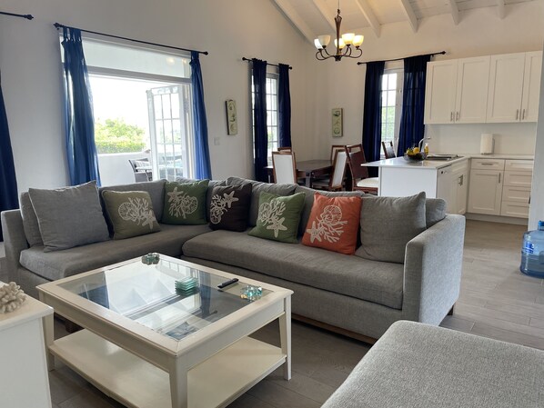 A modern living room with a sectional sofa, a glass coffee table, and a dining area in the background.