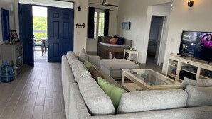 Ein Wohnzimmer mit grauem Sofa, einem Couchtisch und einem Fernseher. Eine blaue Tür führt nach draußen, ein kleiner Tisch mit einer Pflanze und eine weiße Kommode.