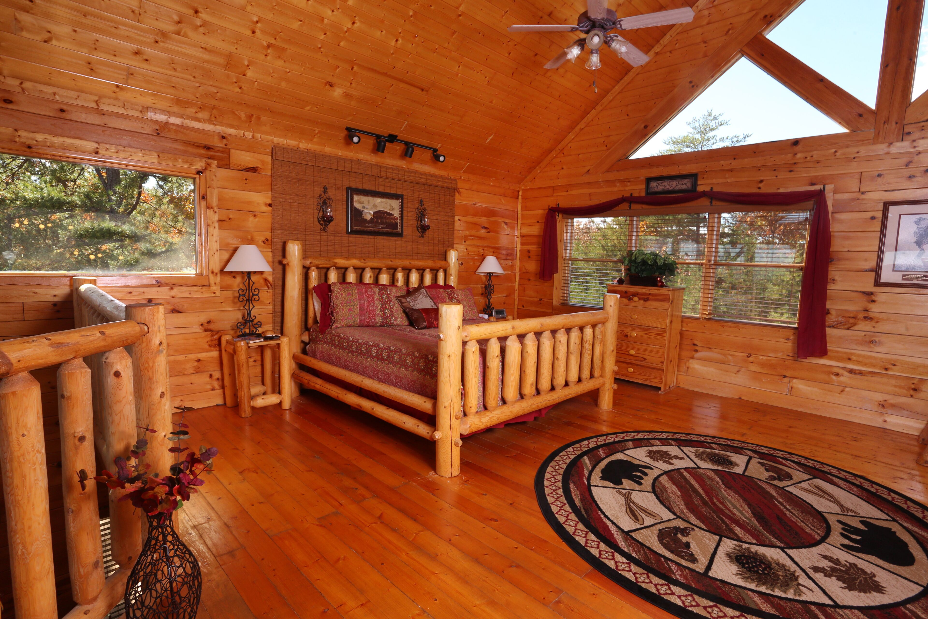A log cabin bedroom with a wooden bed, a nightstand, a lamp, a window with a view of trees, and a ceiling fan.