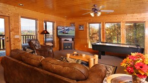 Un intérieur de chalet chaleureux avec un canapé en cuir marron, une table de billard, un téléviseur et une cheminée.