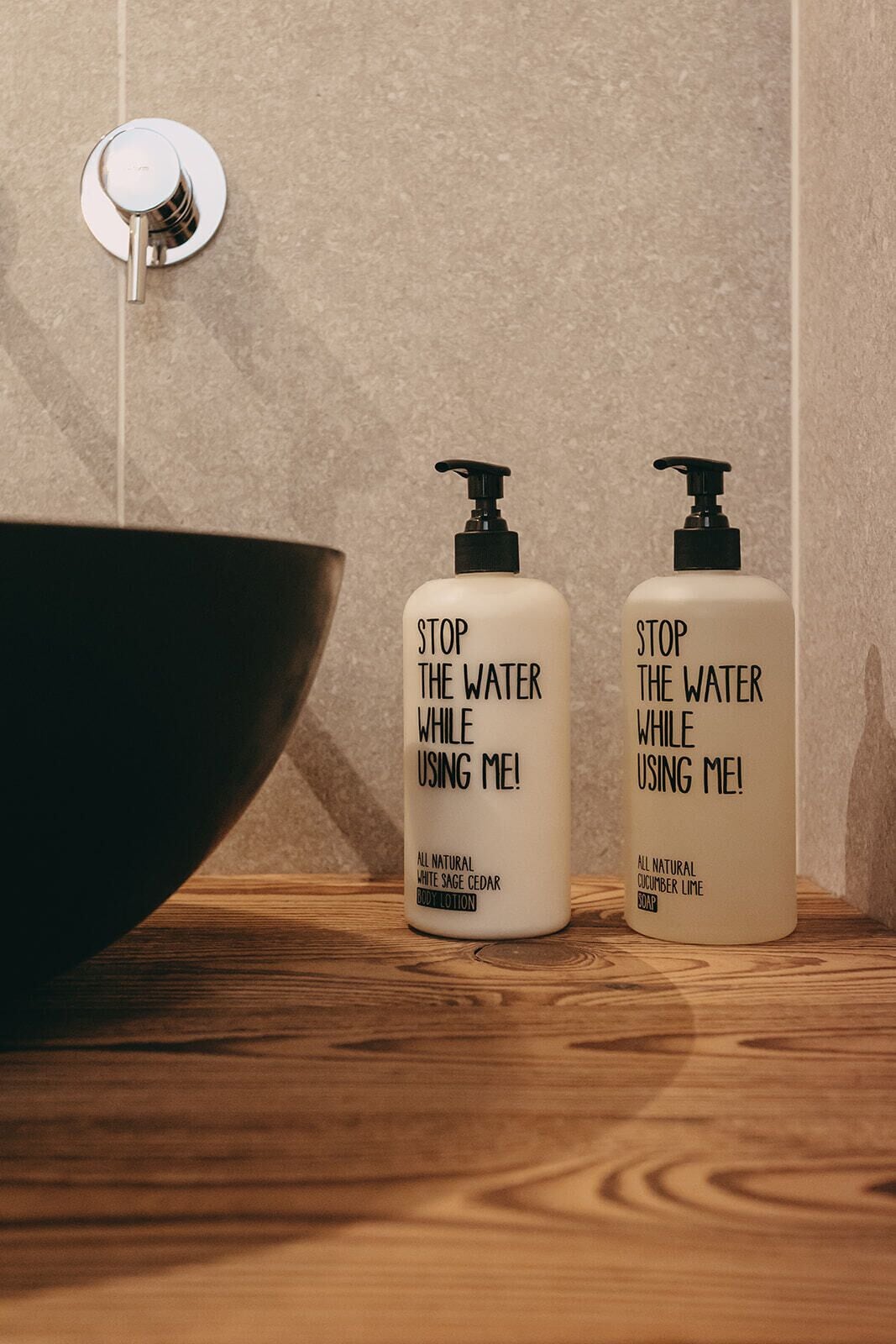 To flasker med bodylotion og sæbe på en træoverflade med en skål og hanefaucet i baggrunden.