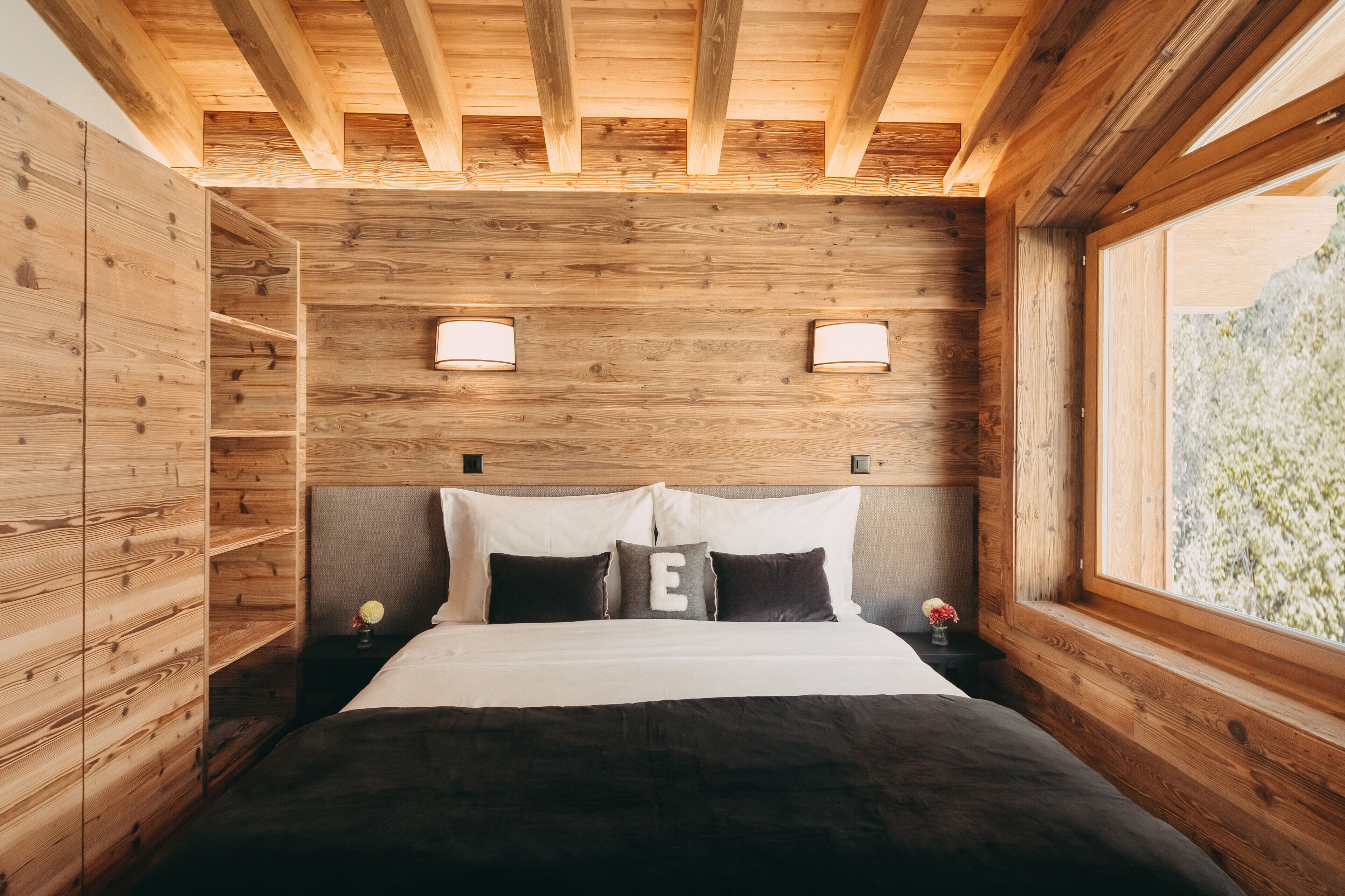 A wooden cabin bedroom with a bed, two bedside lamps, and a window with a view of greenery.
