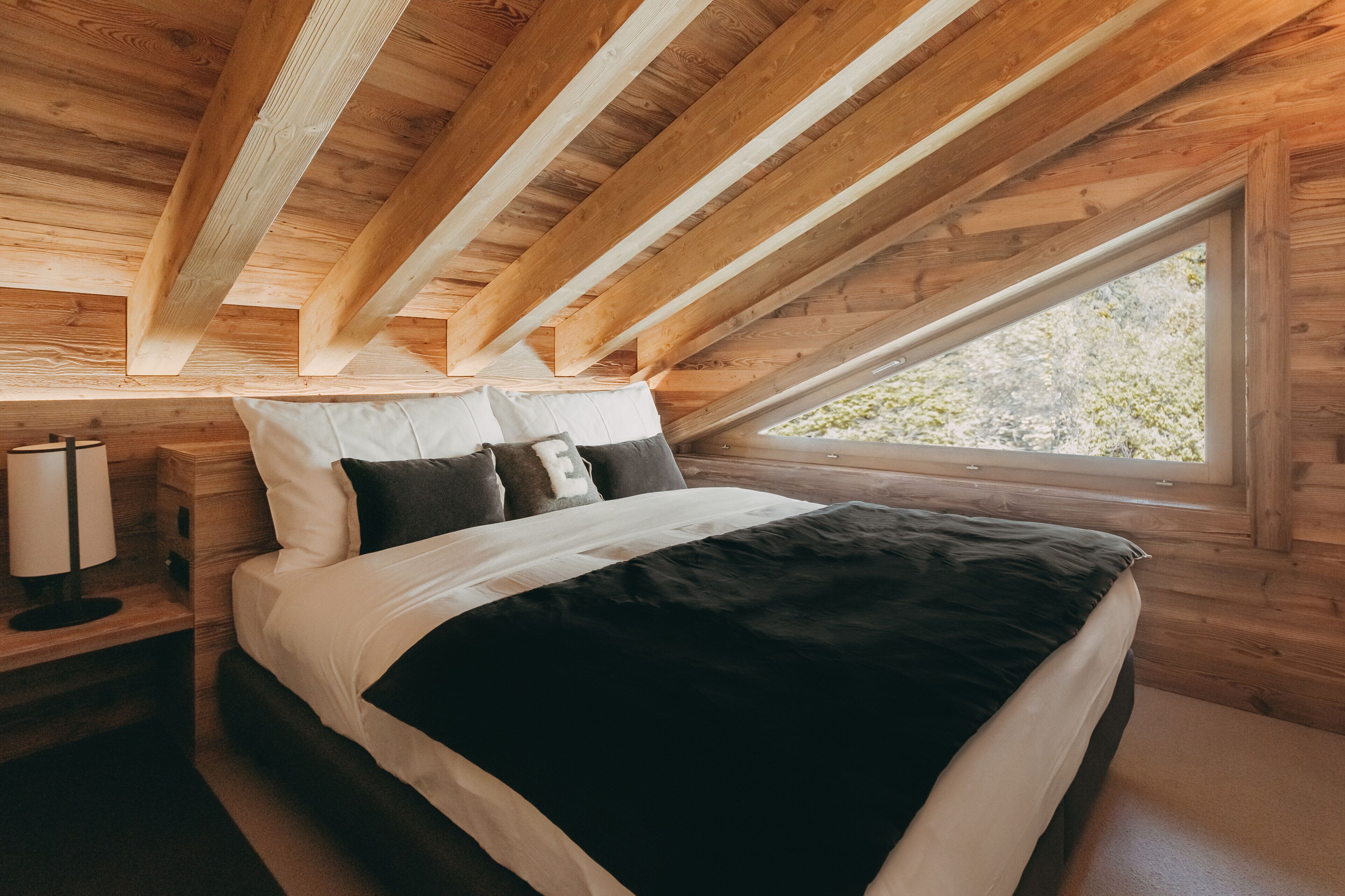 Une chambre avec un plafond en bois, une grande fenêtre, un lit avec une literie blanche et noire, et une table de chevet avec une lampe.
