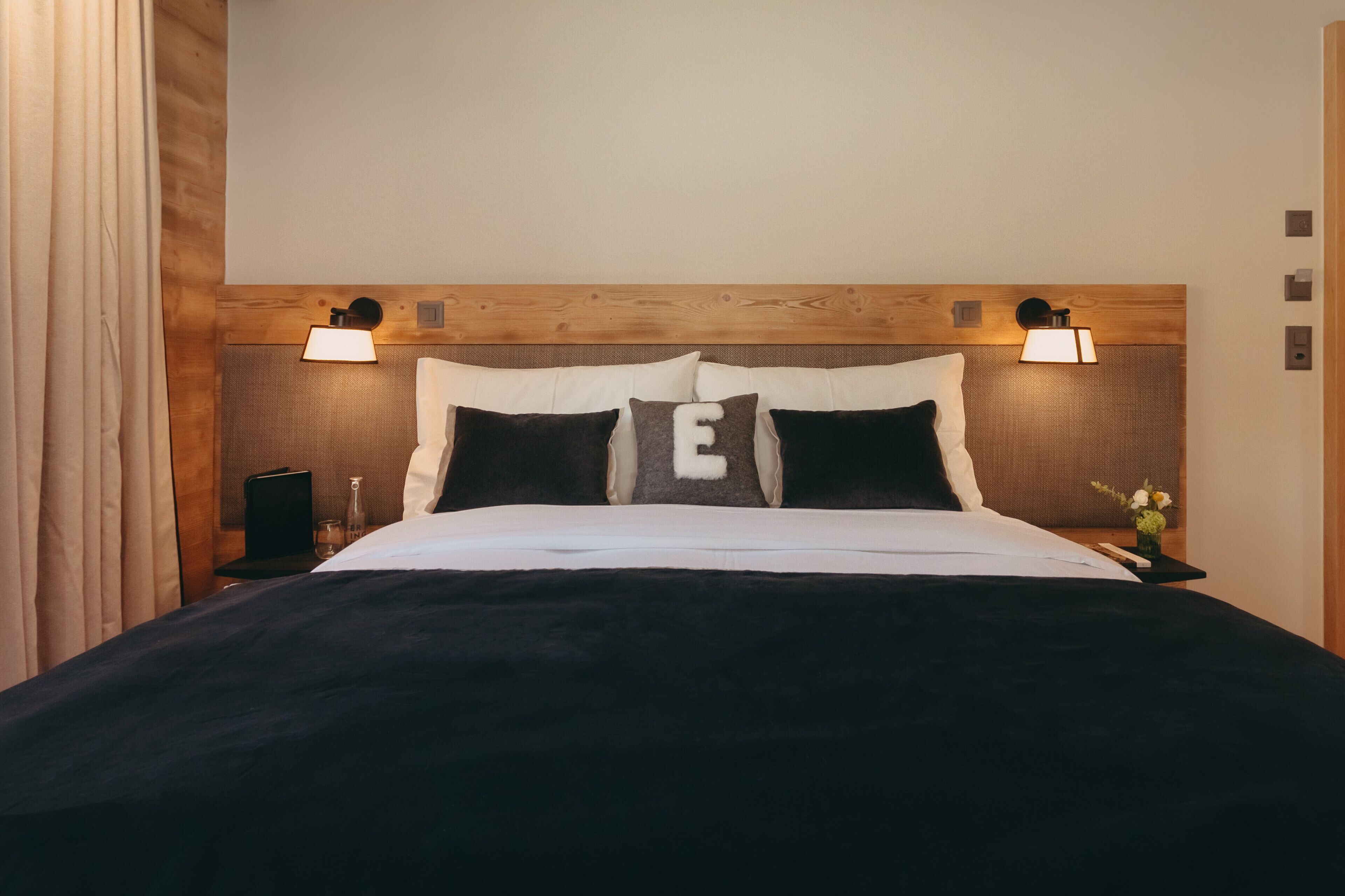 A neatly made bed with a dark duvet, two bedside lamps, and a wooden headboard.