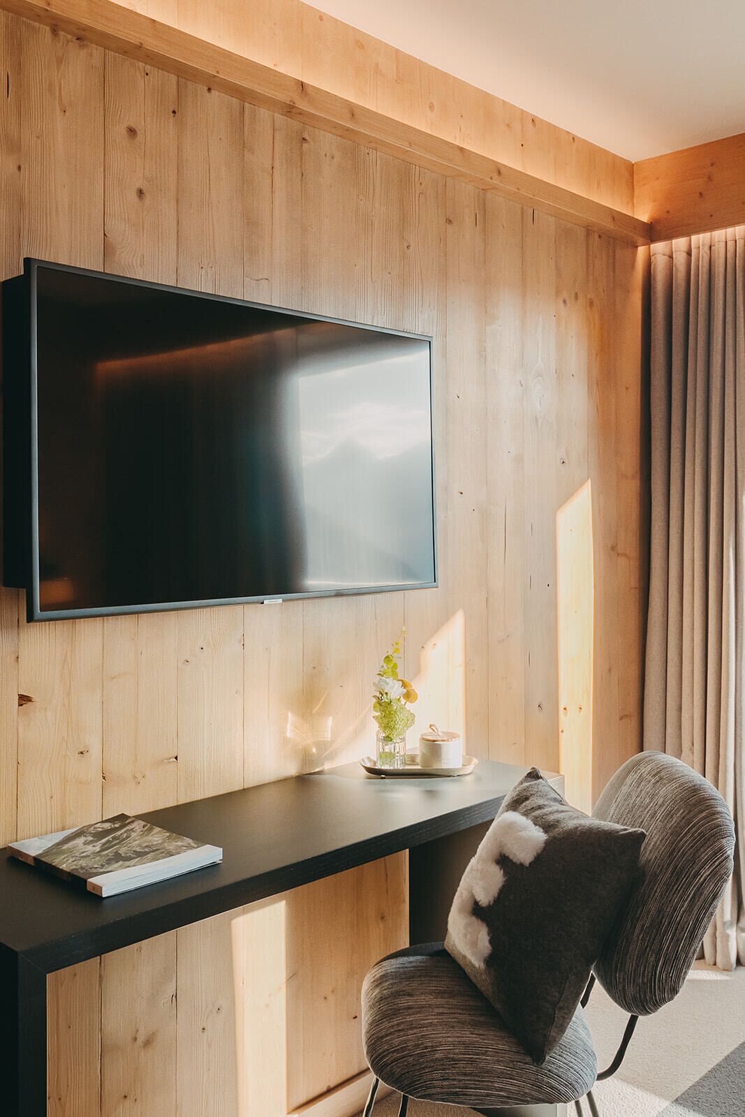 Una camera moderna con parete in legno, una TV a schermo piatto montata sopra una scrivania scura, una sedia con cuscino decorato e un tavolino con un vaso e una tazza.