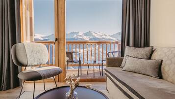 A room with a sofa, two chairs, a small table with a vase, and a view of snow-capped mountains through large windows.