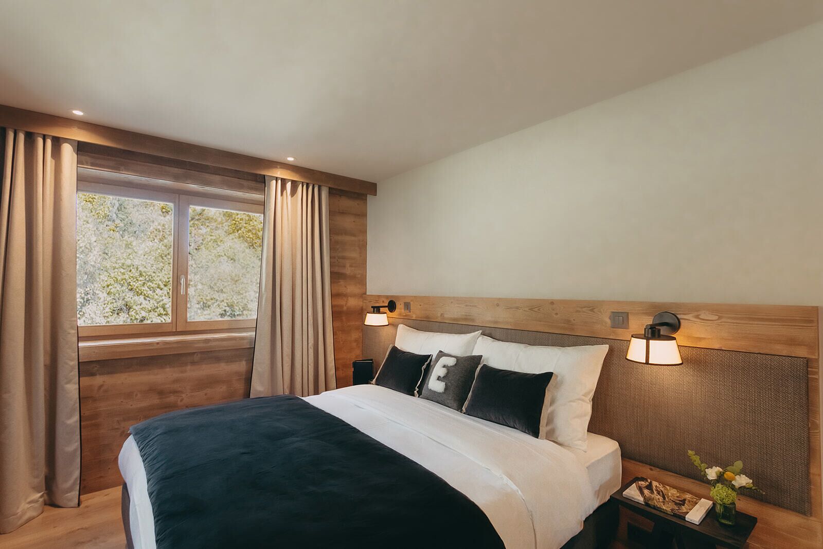 A bedroom with a large bed, wooden headboard, two bedside lamps, a nightstand, and a window with curtains.