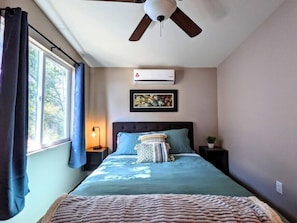 Ein Schlafzimmer mit Bett, Deckenventilator, Klimaanlage, Nachttisch, Lampe, Fenster mit Vorhängen und einem Wandbild.