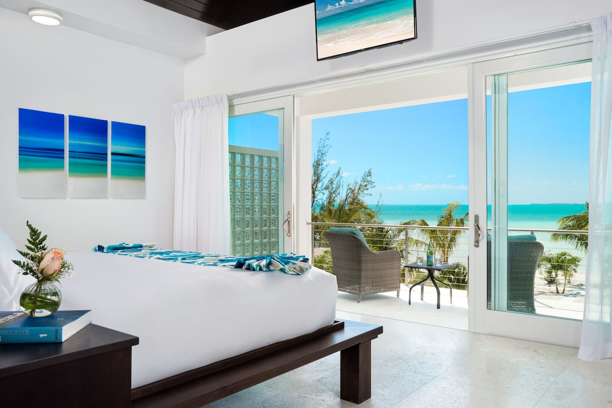 A hotel room with a bed, a bedside table, a TV, and a balcony with a view of the beach and ocean.