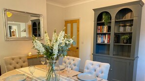 Uma sala de jantar com uma mesa redonda posta para quatro pessoas, um vaso de flores brancas e uma estante com livros e objetos decorativos.