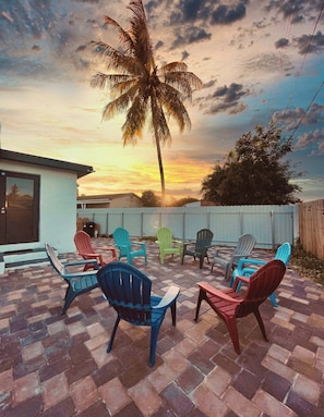 Een terras buiten met een tegelvloer, diverse Adirondack-stoelen en een palmboom.