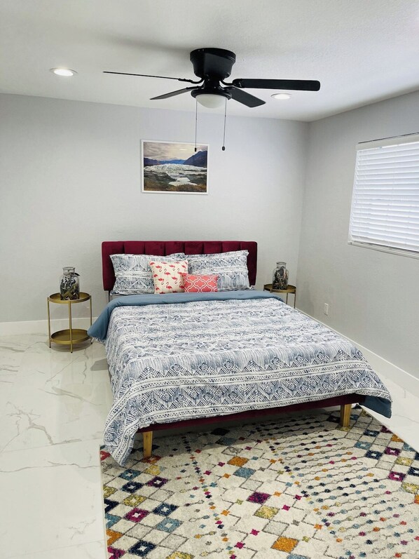 Un dormitorio con cama, ventilador de techo, ventana con persianas, un cuadro en la pared y una alfombra estampada.