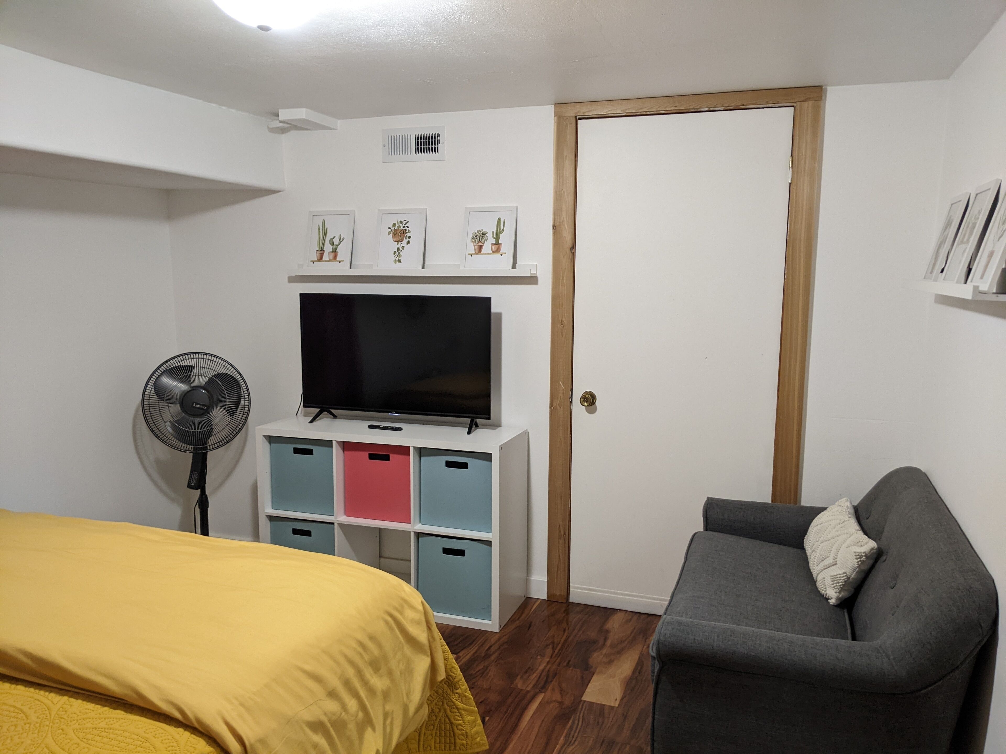 Une chambre avec un lit, un téléviseur posé sur un meuble, un ventilateur et une porte.