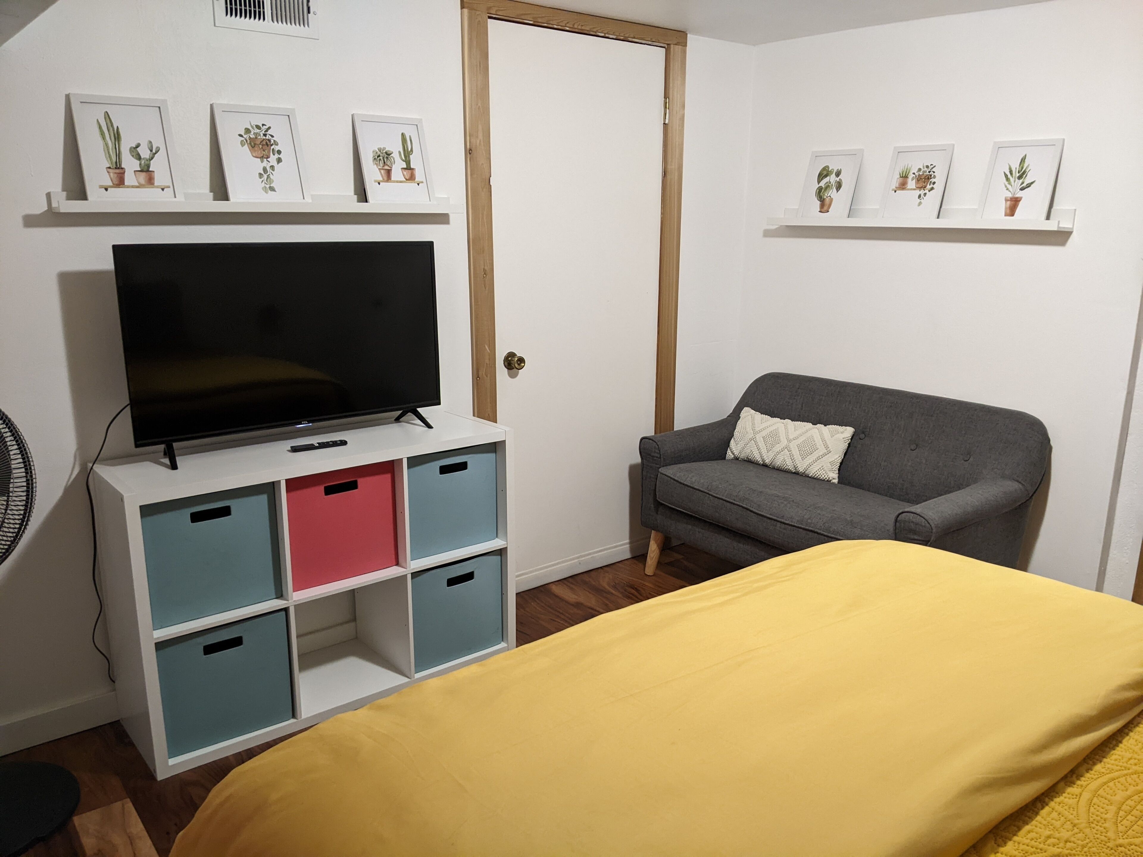 A room with a bed, a TV on a white stand, a grey sofa, and storage units with colored doors.