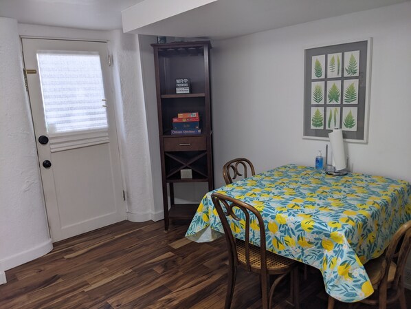 A dining area with a table covered in a floral tablecloth, two wooden chairs, a wooden cabinet with a 'WiFi Password' sign, and a window with blinds.