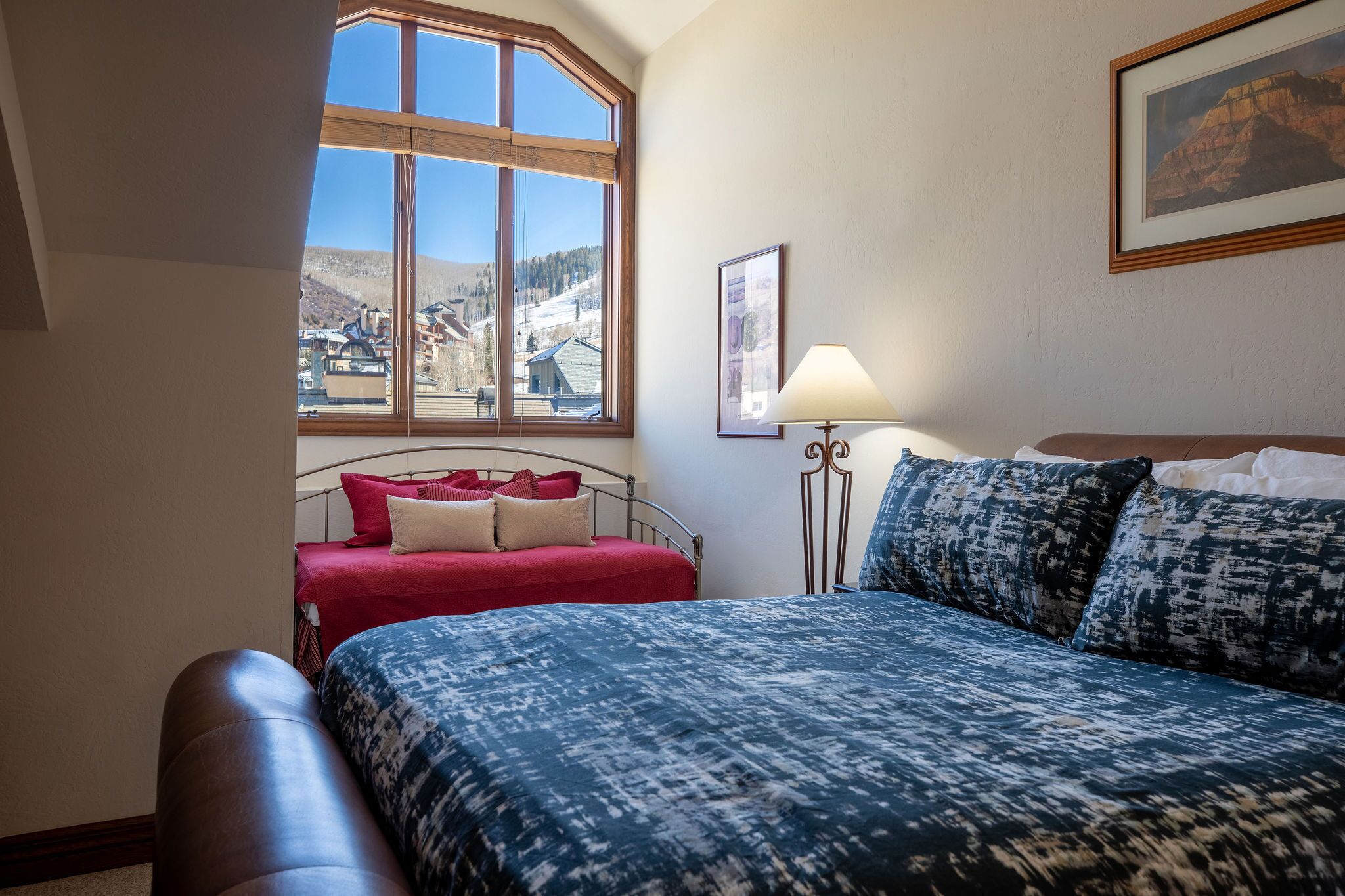 Een slaapkamer met een groot bed, een raam met uitzicht op de bergen, een nachtkastje met een lamp en een ingelijst beeld aan de muur.