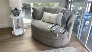 Un canapé d’angle agrémenté d’un plaid, une petite table d’appoint blanche avec une lampe, et une vue sur l’extérieur grâce à une grande fenêtre.