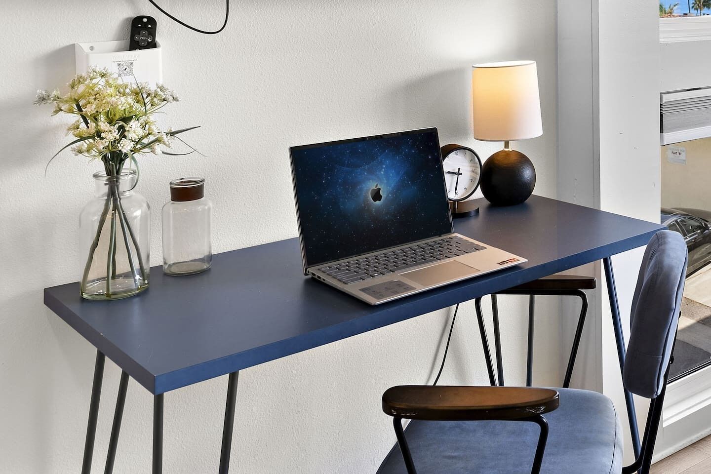 A laptop on a desk with a lamp, a clock, and a vase with flowers.