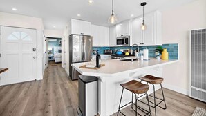 A modern kitchen with white cabinets, stainless steel appliances, a central island with stools, pendant lighting, and a view into the adjacent dining area. - Oceanview Beach Retreat Walk to Pier AC Pets ok (San Clemente)