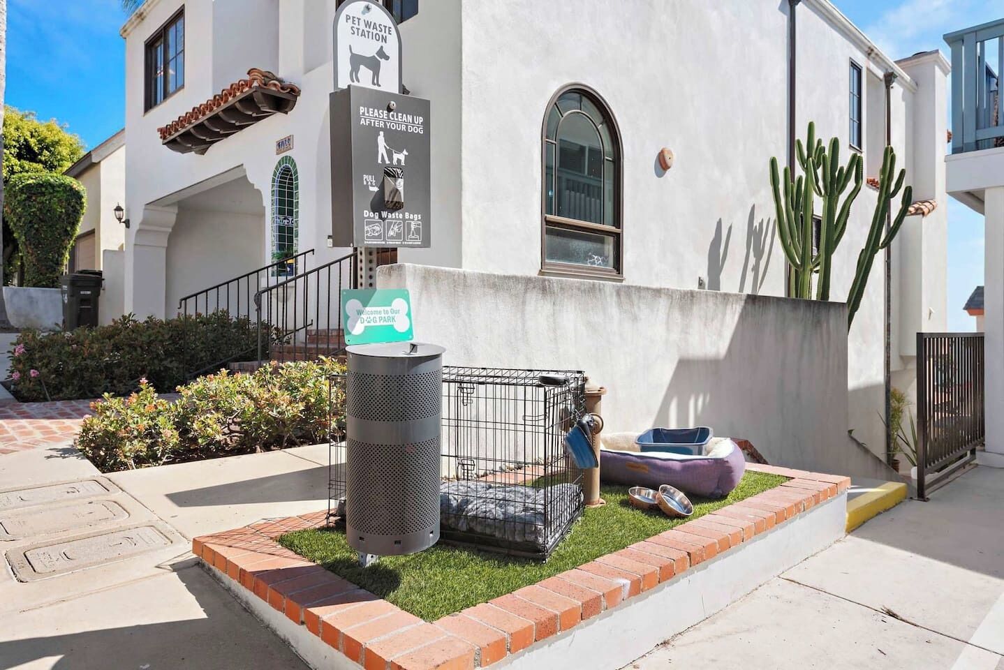 Una estación para desechos de mascotas con un letrero, un área cercada con dispensador de bolsas para excrementos de perro y una casita pequeña para perros.