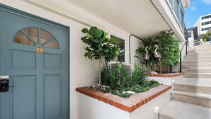 A blue door with a keypad lock, a small garden with various plants, and a concrete staircase leading to a building.