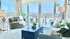 A spacious living room with a large window overlooking the sea, a blue coffee table, and a grey sofa.