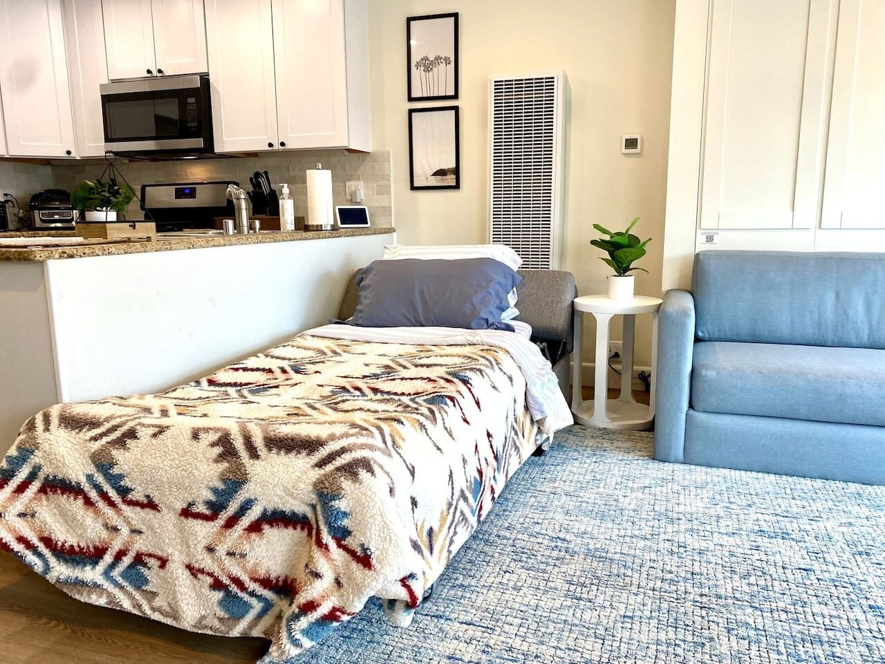 Un dormitorio pequeño con cama, mesita de noche, silla y espacio para kitchenette.