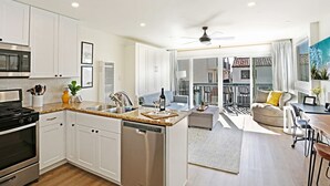 Eine moderne Küche mit weißen Schränken, einer Granit-Arbeitsplatte und Edelstahlgeräten. Es gibt einen Essbereich mit Tisch und Stühlen, eine Couch sowie einen Balkon mit Blick.