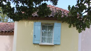 Un bâtiment à deux étages, avec une porte rouge et des volets bleus, entouré de verdure.
